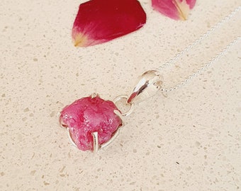 May include: A silver chain necklace with a pendant featuring a raw pink ruby gemstone. The ruby is set in a silver frame and has a rough, natural appearance.