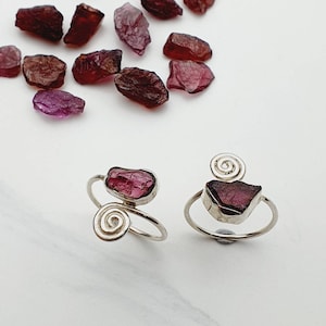 May include: Two silver rings, each with a raw, dark pink gemstone. One ring has a spiral design, the other a spiral accent. Several raw gemstones in shades of pink and purple are scattered around the rings.