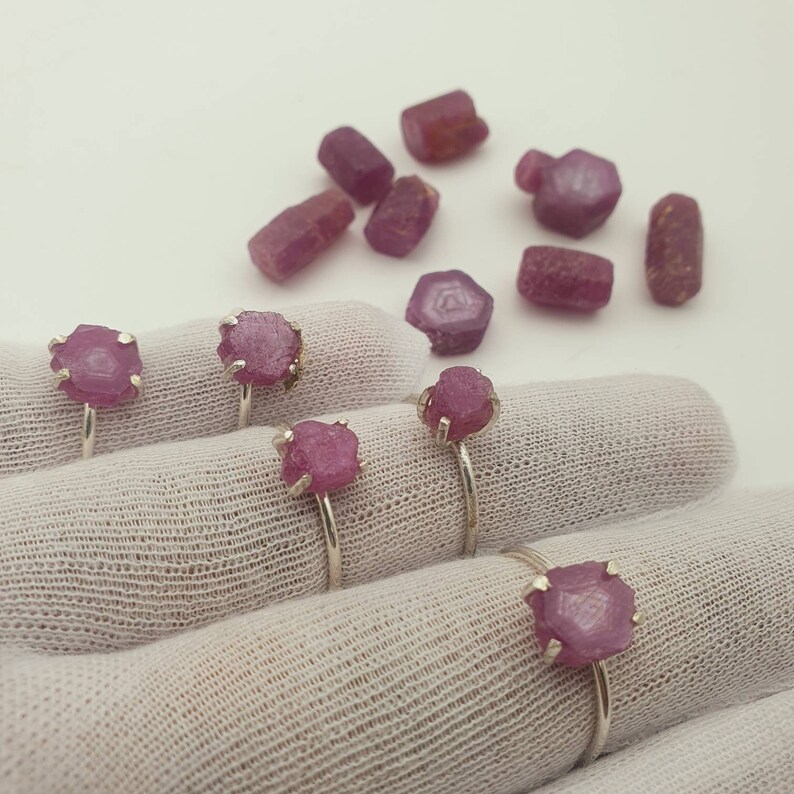May include: Five silver rings with pink ruby gemstones set in the center of each ring. The rings are displayed on a white fabric surface.
