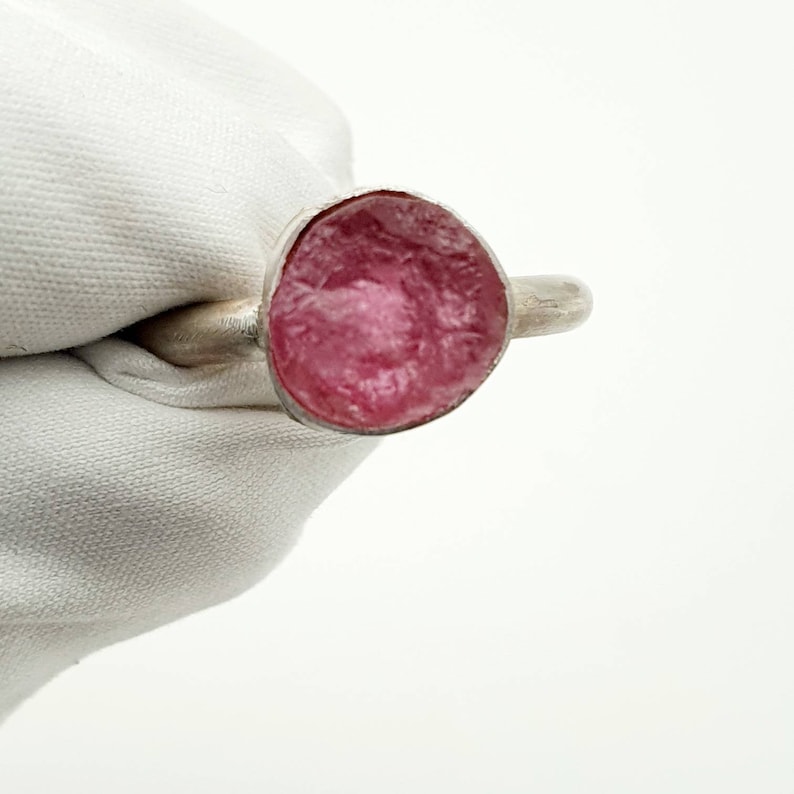 Op de afbeelding: Een zilveren ring met een grote, ronde, roze edelsteen. De edelsteen heeft een textuur en is gezet in een eenvoudige zilveren band. De ring wordt getoond tegen een witte achtergrond.