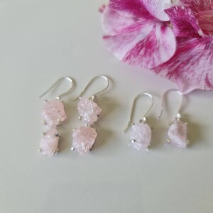 May include: A pair of silver earrings with two pink crystal drops each. The earrings are on a white surface with a pink flower in the background.
