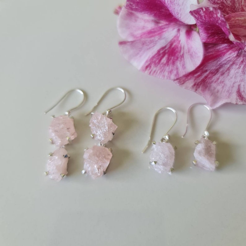 May include: A pair of silver earrings with two pink crystal drops each. The earrings are on a white surface with a pink flower in the background.