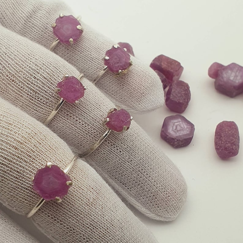 May include: Five silver rings with pink gemstone settings. The rings are displayed on white fabric. Loose pink gemstones are scattered around the rings.
