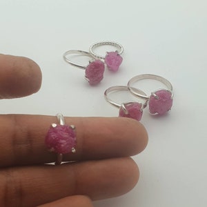 Peut inclure: Quatre bagues en argent avec des pierres pr&eacute;cieuses en rubis brut rose. Les bagues sont sur une surface blanche et un doigt porte l'une des bagues.