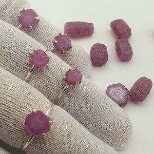 May include: Three silver rings with pink rough cut gemstone settings. The rings are displayed on a white fabric surface. There are several loose pink gemstones scattered around the rings.
