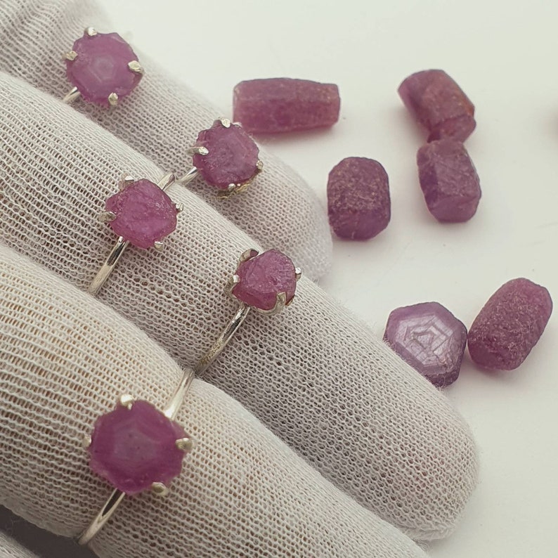 May include: Three silver rings with pink rough cut gemstone settings. The rings are displayed on a white fabric surface. There are several loose pink gemstones scattered around the rings.