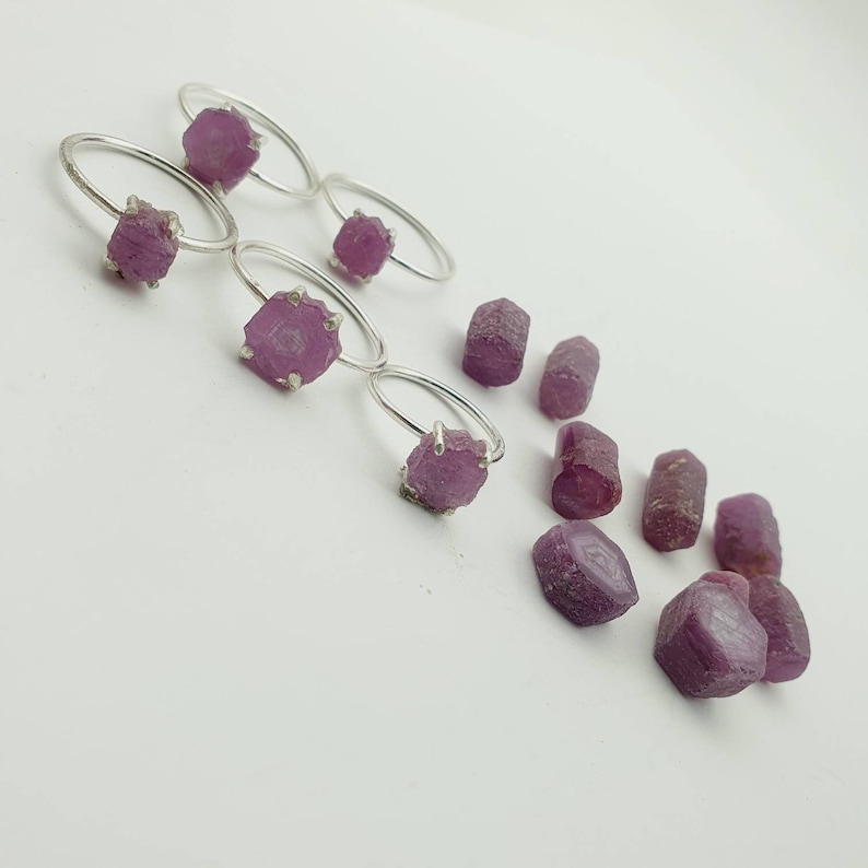 May include: A set of silver rings with purple gemstone settings. The rings are arranged in a row, with the gemstones facing up. There are also several loose purple gemstones scattered around the rings.