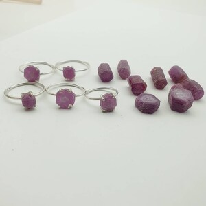 May include: A set of silver rings with pink rough cut gemstones. The rings are arranged in a row with a few loose gemstones scattered around them.