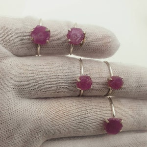 May include: Five silver rings with pink gemstone settings. The rings are stacked on top of each other on a white fabric surface.