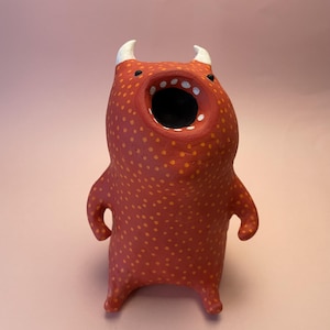 Può includere: Figurina di mostro in ceramica fatta a mano. Il corpo rosso è macchiato di punti arancioni, con corna bianche e una bocca spalancata. La figurina ha braccia e gambe piccole e sta in piedi. Il design è giocoso e unico.