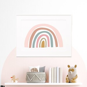 May include: A white bed with a brown and white striped blanket and a yellow throw blanket. There are several pillows on the bed, including a brown and white striped pillow, a gray pillow with a rabbit design, and a brown pillow with a fringe. There is a white nightstand with a lamp and a small wooden chair with white bunny ears. A framed print of a rainbow with pink, orange, yellow, green, and blue colors hangs on the wall above the bed.