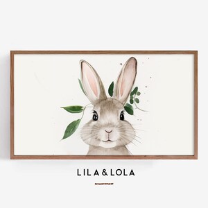 May include: Watercolor print of a bunny with long ears and green leaves. The bunny is light brown with pink inner ears and is set against a white background. The print is in a wooden frame. The text "LILA & LOLA" is at the bottom.