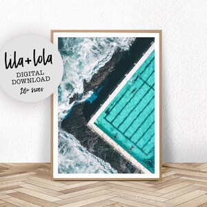 May include: An aerial view of a swimming pool with turquoise water and white lines. The pool is surrounded by dark rocks and crashing waves.