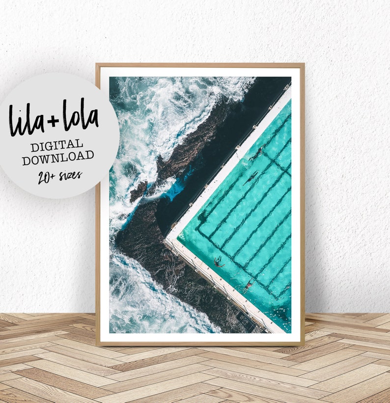 May include: An aerial view of a swimming pool with turquoise water and white lines. The pool is surrounded by dark rocks and crashing waves.
