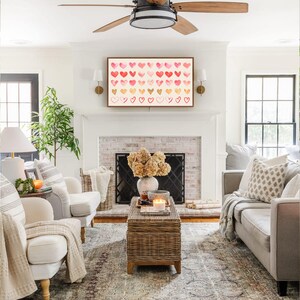May include: A living room with a white brick fireplace, a white couch, two white armchairs, a coffee table, and a framed print of a watercolor painting of pink and red hearts.