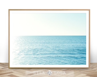May include: A framed print featuring a serene ocean scene. The artwork displays a clear blue sky meeting the calm, turquoise water at the horizon. The print is encased in a light brown wooden frame, with the text "LILA LOLA" at the bottom.