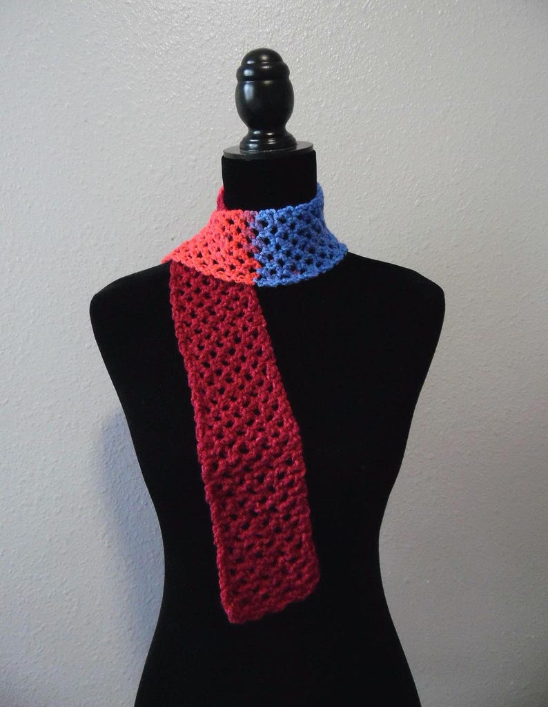 May include: A handmade crochet scarf in a color-block design. The scarf features a red section, transitioning to a pink section, and then to a blue section. The scarf is displayed on a black mannequin, showcasing its open weave and color scheme.