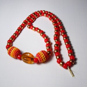 May include: A red and gold beaded necklace with crocheted accents and a clear amber bead.