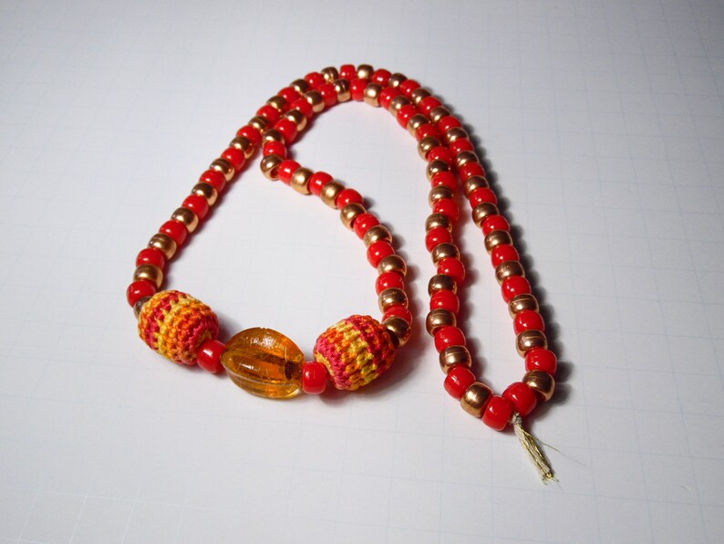 May include: A red and gold beaded necklace with crocheted accents and a clear amber bead.