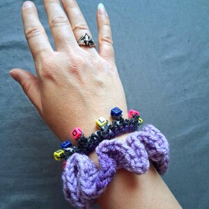 K&ouml;nnte beinhalten: Nahaufnahme einer lila H&auml;kel-Haargummi mit einem schwarz-grauen H&auml;kelband, das mit bunten W&uuml;rfelperlen verziert ist, die Buchstaben buchstabieren. Ein silberner Ring mit einem dunklen Stein befindet sich an einer Hand.