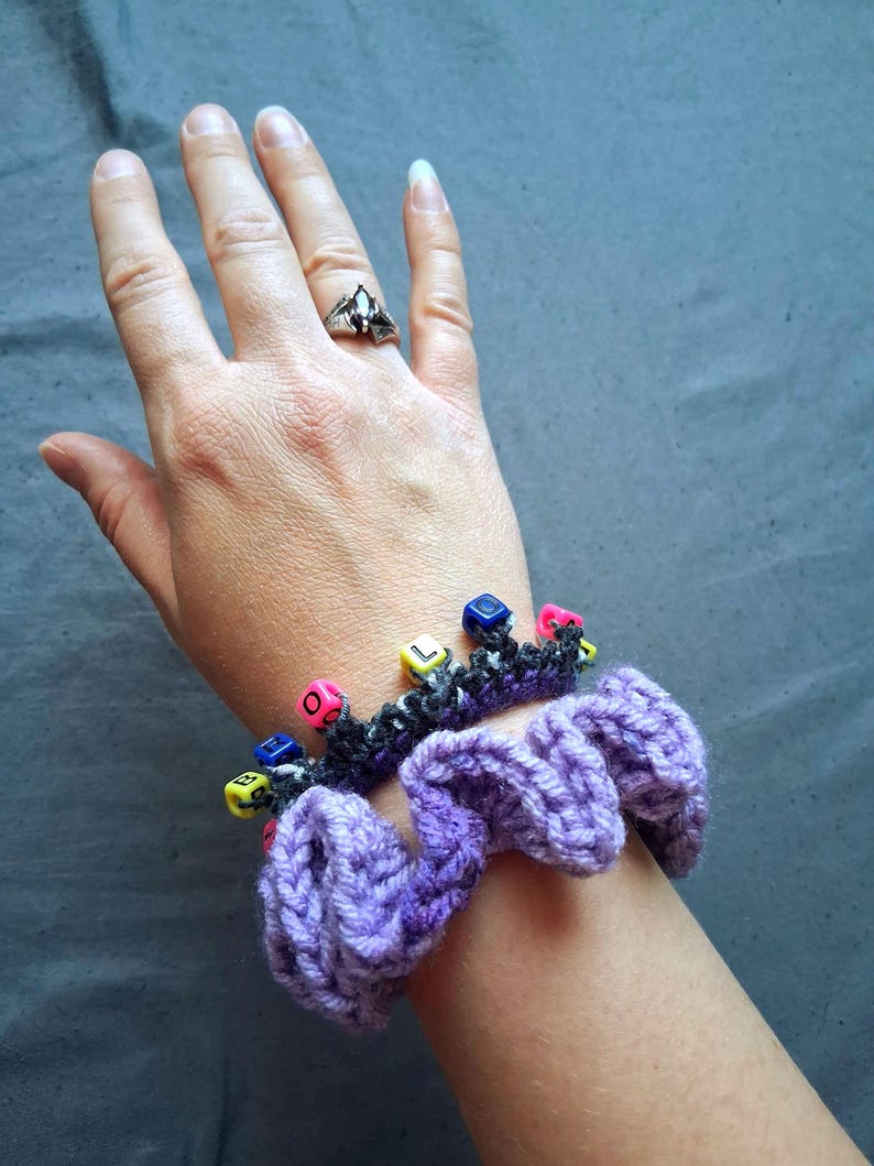 K&ouml;nnte beinhalten: Nahaufnahme einer lila H&auml;kel-Haargummi mit einem schwarz-grauen H&auml;kelband, das mit bunten W&uuml;rfelperlen verziert ist, die Buchstaben buchstabieren. Ein silberner Ring mit einem dunklen Stein befindet sich an einer Hand.