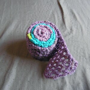 May include: A rolled-up, handmade crochet scarf in shades of purple, teal, and pink. The scarf has a spiral design with a teal center and a decorative, openwork edge. The item is displayed on a gray surface.
