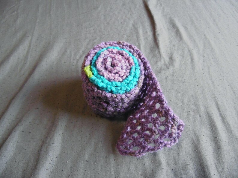 May include: A rolled-up, handmade crochet scarf in shades of purple, teal, and pink. The scarf has a spiral design with a teal center and a decorative, openwork edge. The item is displayed on a gray surface.