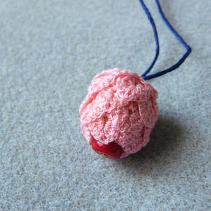Puede incluir: Un colgante rosa hecho a mano con ganchillo y una pequeña cuenta roja en la parte inferior, suspendido de un cordón azul. El colgante tiene una forma redondeada y texturizada. El fondo es de tela gris claro.
