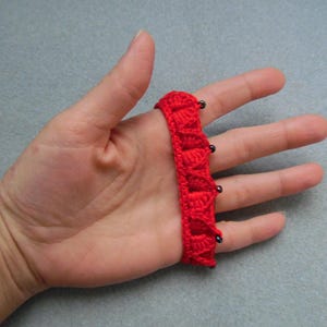 May include: Red crocheted fingerless gloves with black beads. The gloves are worn on a hand.