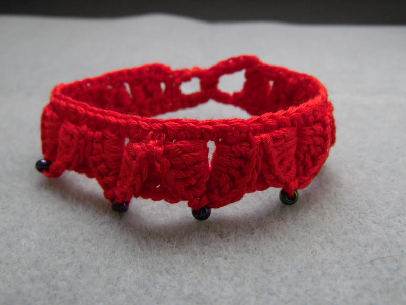 May include: A red crocheted bracelet with black beads. The bracelet is made with a simple stitch and has a delicate, feminine look.
