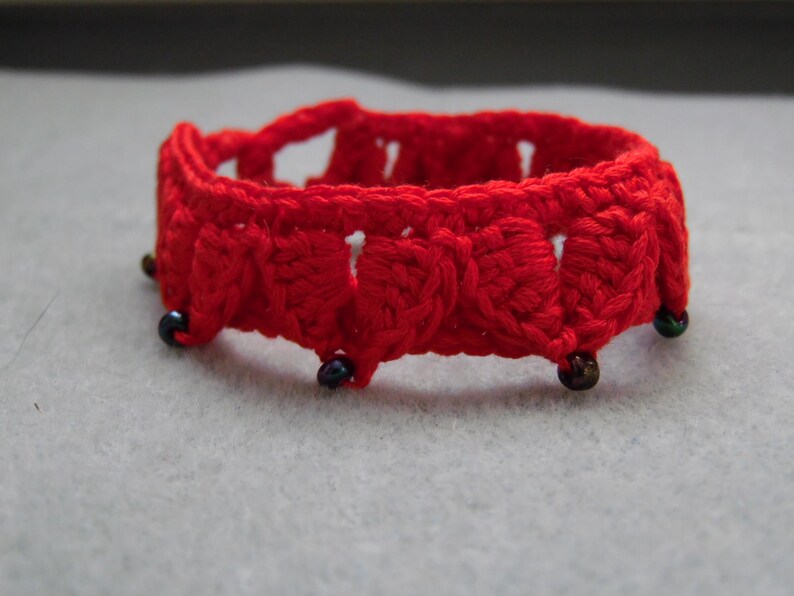 May include: Red crocheted bracelet with black beads. The bracelet is made of a delicate, lacy pattern.