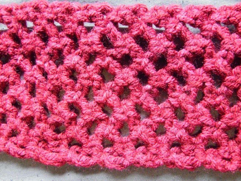 May include: Close-up of a section of hand-crocheted fabric in a vibrant shade of red. The openwork design creates a lacy pattern with visible holes. The texture of the yarn is clearly visible, showcasing the handmade quality of the textile.