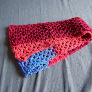 May include: A handmade, crocheted cowl in shades of red, coral, and blue. The cowl is folded, revealing the different color sections. The crochet stitch creates an open, textured pattern. This accessory is suitable for cold weather.