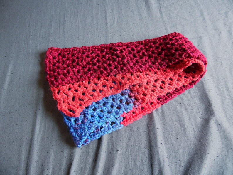 May include: A handmade, crocheted cowl in shades of red, coral, and blue. The cowl is folded, revealing the different color sections. The crochet stitch creates an open, textured pattern. This accessory is suitable for cold weather.