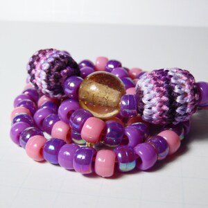 Puede incluir: Un collar de cuentas de color morado y rosa con dos cuentas de ganchillo y una cuenta de vidrio transparente.