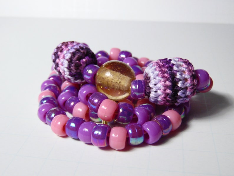 Puede incluir: Un collar de cuentas de color morado y rosa con dos cuentas de ganchillo y una cuenta de vidrio transparente.