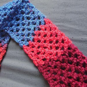 May include: A handmade crochet scarf with a gradient of colors. The scarf transitions from blue to red and then to a deep burgundy. The open weave design is visible, creating a textured appearance. The scarf is likely made of soft yarn.