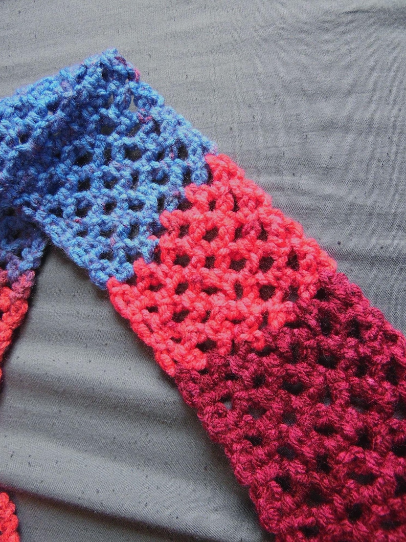May include: A handmade crochet scarf with a gradient of colors. The scarf transitions from blue to red and then to a deep burgundy. The open weave design is visible, creating a textured appearance. The scarf is likely made of soft yarn.