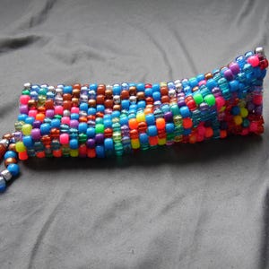 May include: A colorful beaded pouch with a wrist strap. The pouch is made of small, round beads in various colors, including blue, pink, orange, and brown. The wrist strap is made of smaller beads in shades of brown and blue.