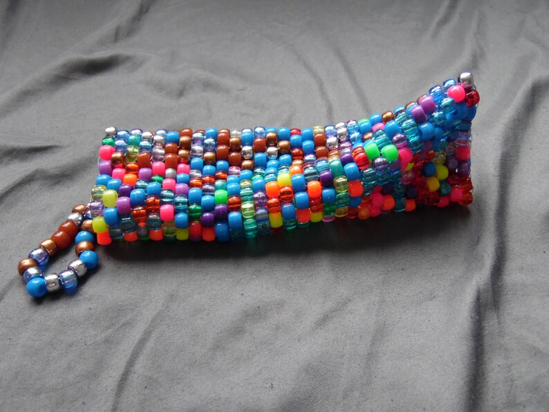 May include: A colorful beaded pouch with a wrist strap. The pouch is made of small, round beads in various colors, including blue, pink, orange, and brown. The wrist strap is made of smaller beads in shades of brown and blue.