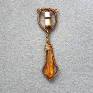May include: A handcrafted pendant with a copper wire frame. The pendant features a central teardrop-shaped element with orange and yellow hues, and two rectangular beads. The wirework is intricate and detailed.