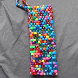 May include: A rectangular beaded pouch in a vibrant array of colors, including blue, orange, pink, green, and purple. The pouch has a loop made of smaller beads at the top. The beads are arranged in a tight, textured pattern.