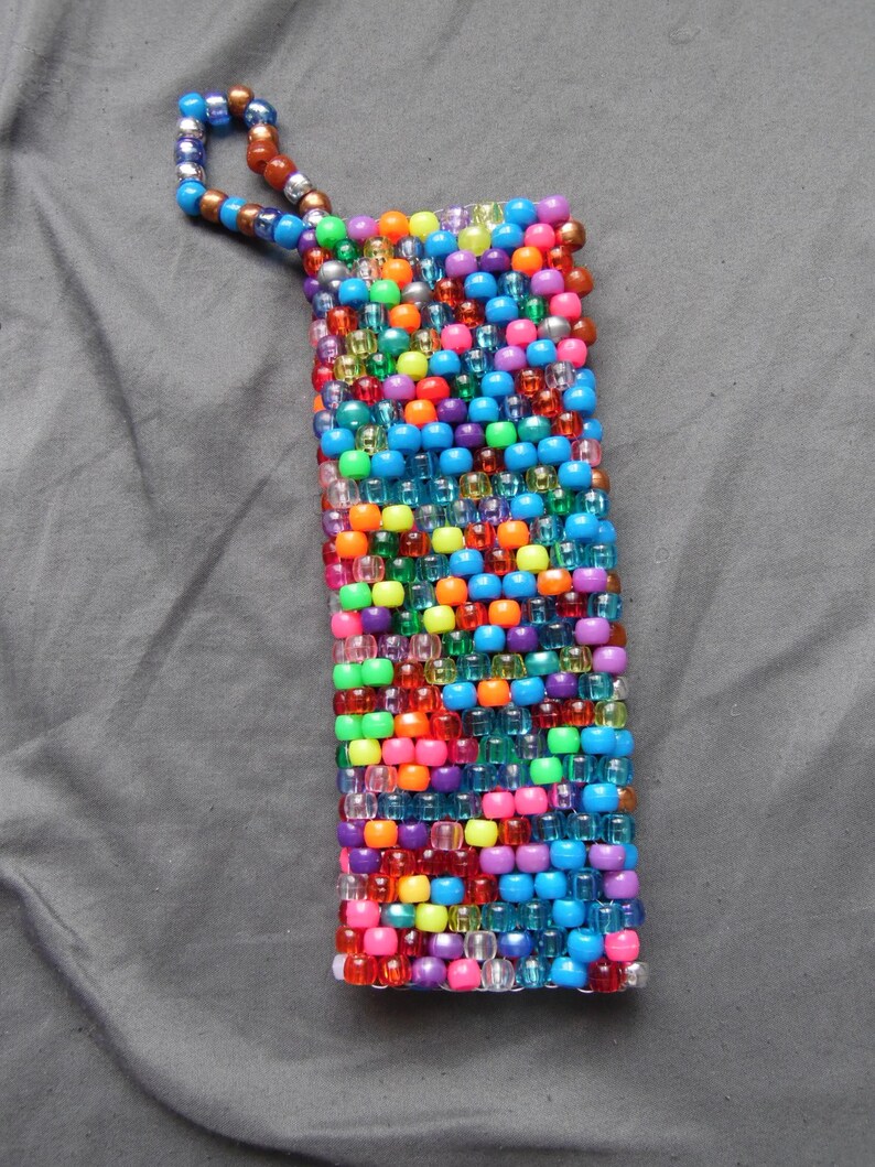 May include: A rectangular beaded pouch in a vibrant array of colors, including blue, orange, pink, green, and purple. The pouch has a loop made of smaller beads at the top. The beads are arranged in a tight, textured pattern.