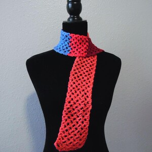 May include: A handmade crochet scarf in a vibrant color block design. The scarf features a blue section at the neck transitioning to a red body. The open weave construction adds a delicate texture. The scarf is displayed on a black mannequin.