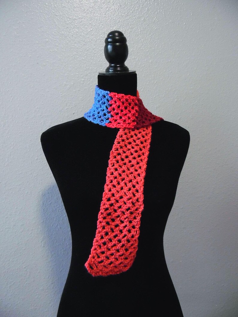 May include: A handmade crochet scarf in a vibrant color block design. The scarf features a blue section at the neck transitioning to a red body. The open weave construction adds a delicate texture. The scarf is displayed on a black mannequin.