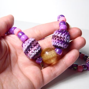 Puede incluir: Un collar de cuentas de color morado y rosa con cuentas de ganchillo y de vidrio. El collar presenta una gran cuenta redonda de vidrio de color &aacute;mbar.