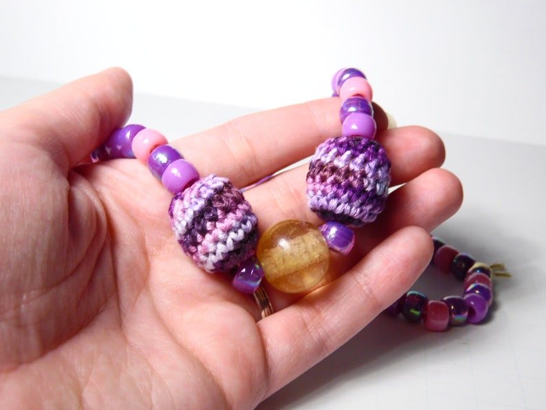 Puede incluir: Un collar de cuentas de color morado y rosa con cuentas de ganchillo y de vidrio. El collar presenta una gran cuenta redonda de vidrio de color &aacute;mbar.