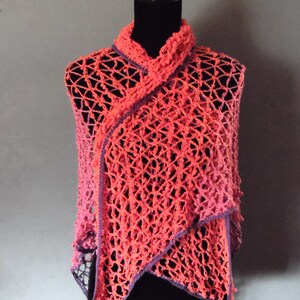 May include: A handmade, open-weave shawl in shades of coral and pink, trimmed with purple. The shawl is draped over a black mannequin, showcasing its asymmetrical design and textured crochet pattern. The garment has a draped collar.