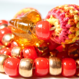 May include: A close-up of a multi-strand necklace with red, gold, and orange beads. The necklace features a crocheted bead with a red, orange, and yellow pattern.