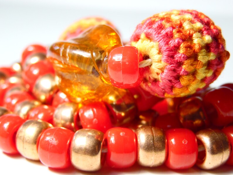 May include: A close-up of a multi-strand necklace with red, gold, and orange beads. The necklace features a crocheted bead with a red, orange, and yellow pattern.